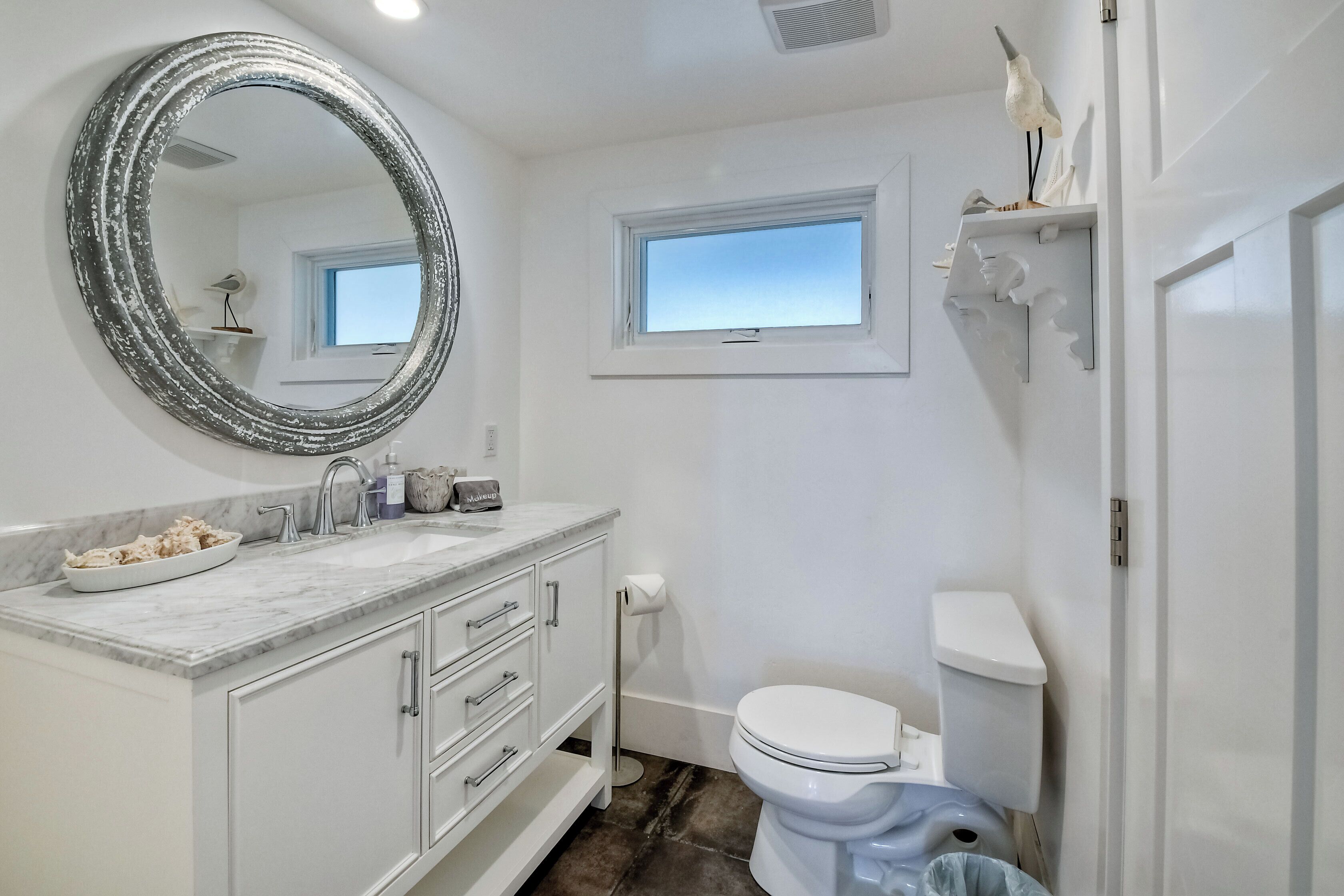 Een badkamer met een wit wastafelmeubel, een ronde spiegel, een toilet en een raam.
