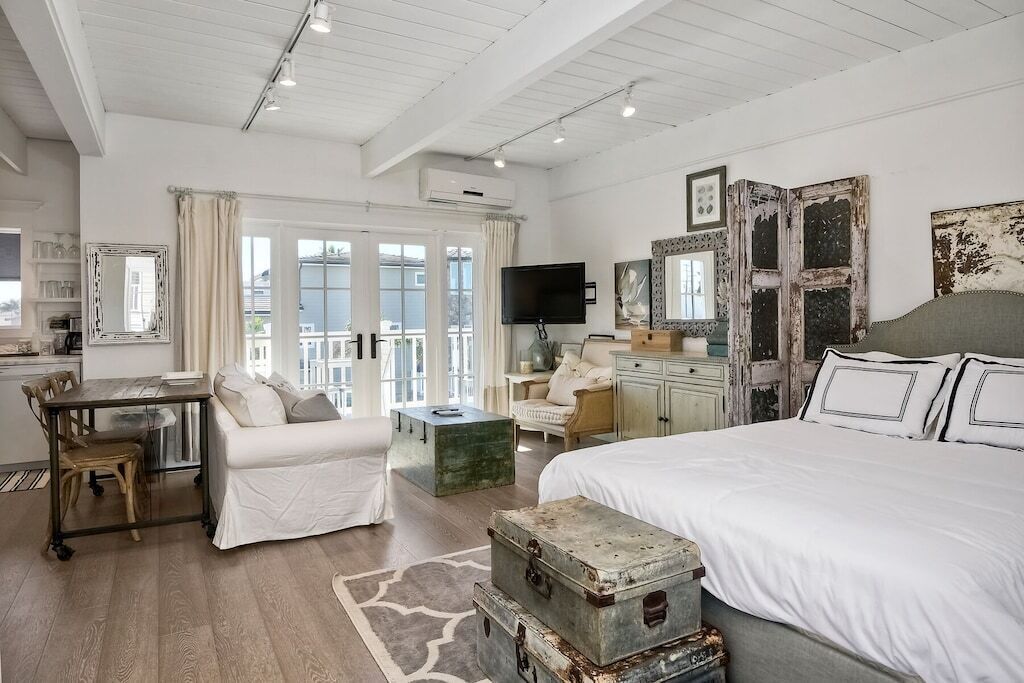 A spacious bedroom with a large bed, a desk, and a chair. There is a balcony visible through the window.