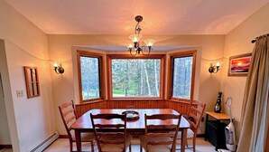 Un comedor con una mesa y sillas de madera, ventanas amplias con vistas a árboles, una araña y una aspiradora.