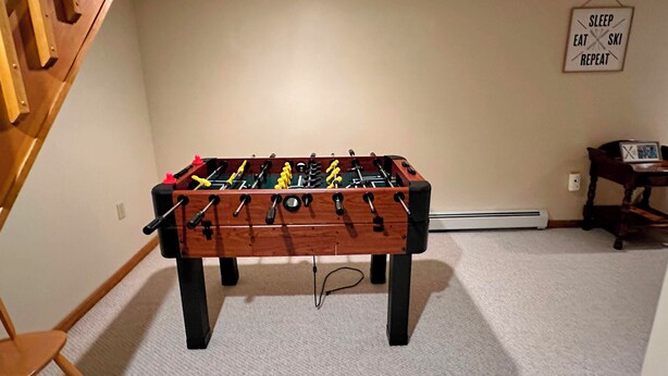 Una habitación con mesa de futbolín, una silla, una mesita con un marco de fotos y un letrero en la pared.