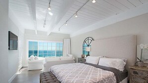 Un espacioso dormitorio con una cama grande, un área de descanso con sofá y sillón, vista al océano a través de amplias ventanas y una mesita de noche de madera rústica.