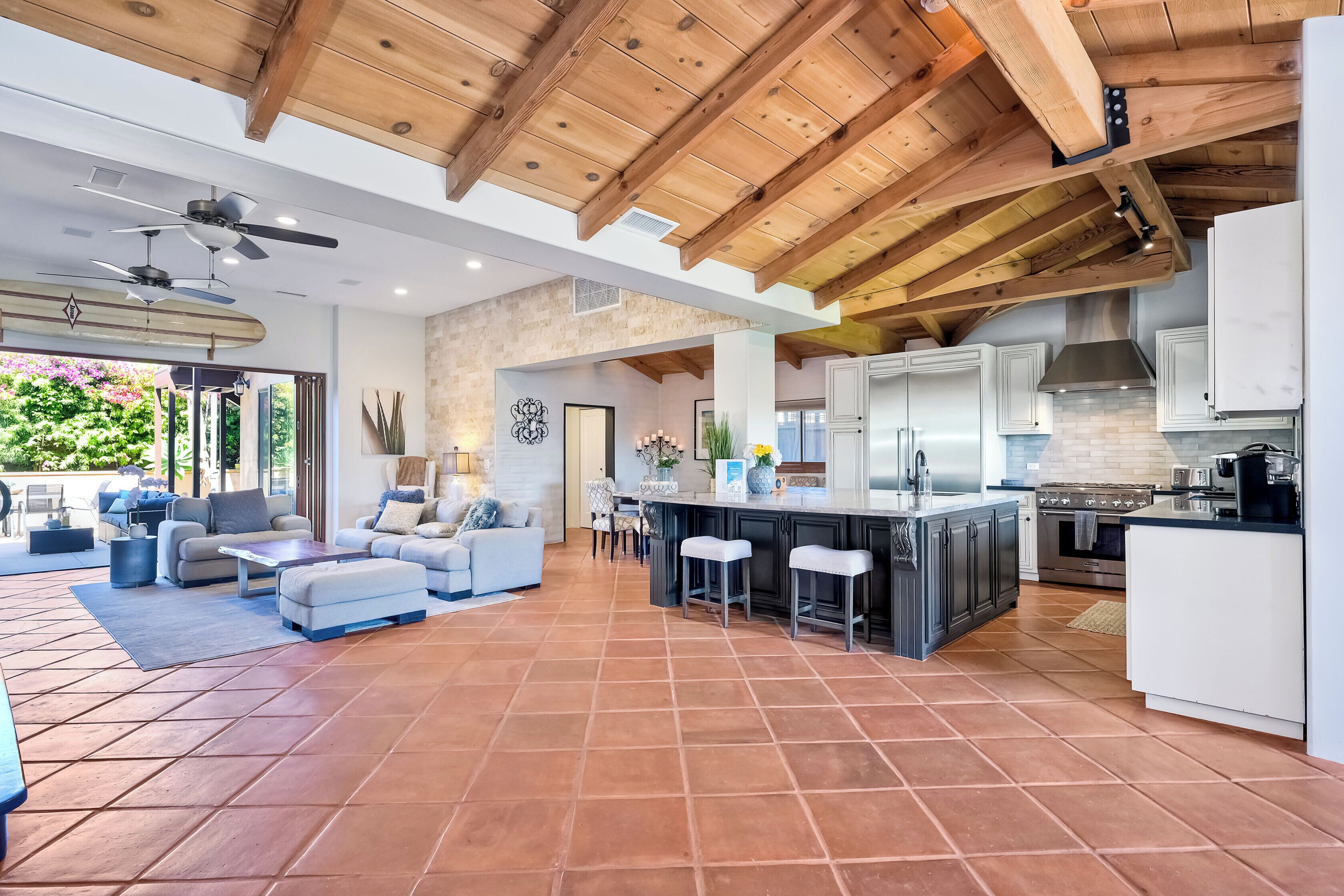 A spacious living area with a high ceiling and wooden beams, a kitchen with modern appliances, and a patio with outdoor furniture.