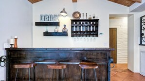 Een bar met een rustieke houten toog en drie krukken. Boven de toog staan decoratieve items, zoals een spiegel, kaarsen en een bordje met de tekst 'DRINK'.