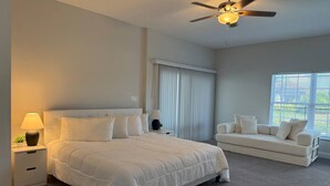 Une chambre à coucher comprenant un lit, des tables de chevet, un ventilateur de plafond, une fenêtre avec des stores et un canapé.