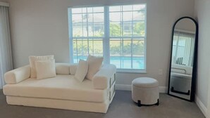 Une pièce spacieuse avec une grande fenêtre, un canapé blanc, un petit pouf et un miroir.