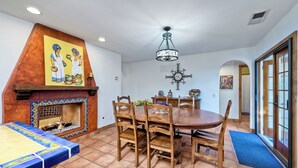 A dining area with a wooden table and chairs, a tiled floor, a fireplace with a decorative tile border, and a ceiling light fixture. - 5100 El Arbol (Carlsbad)