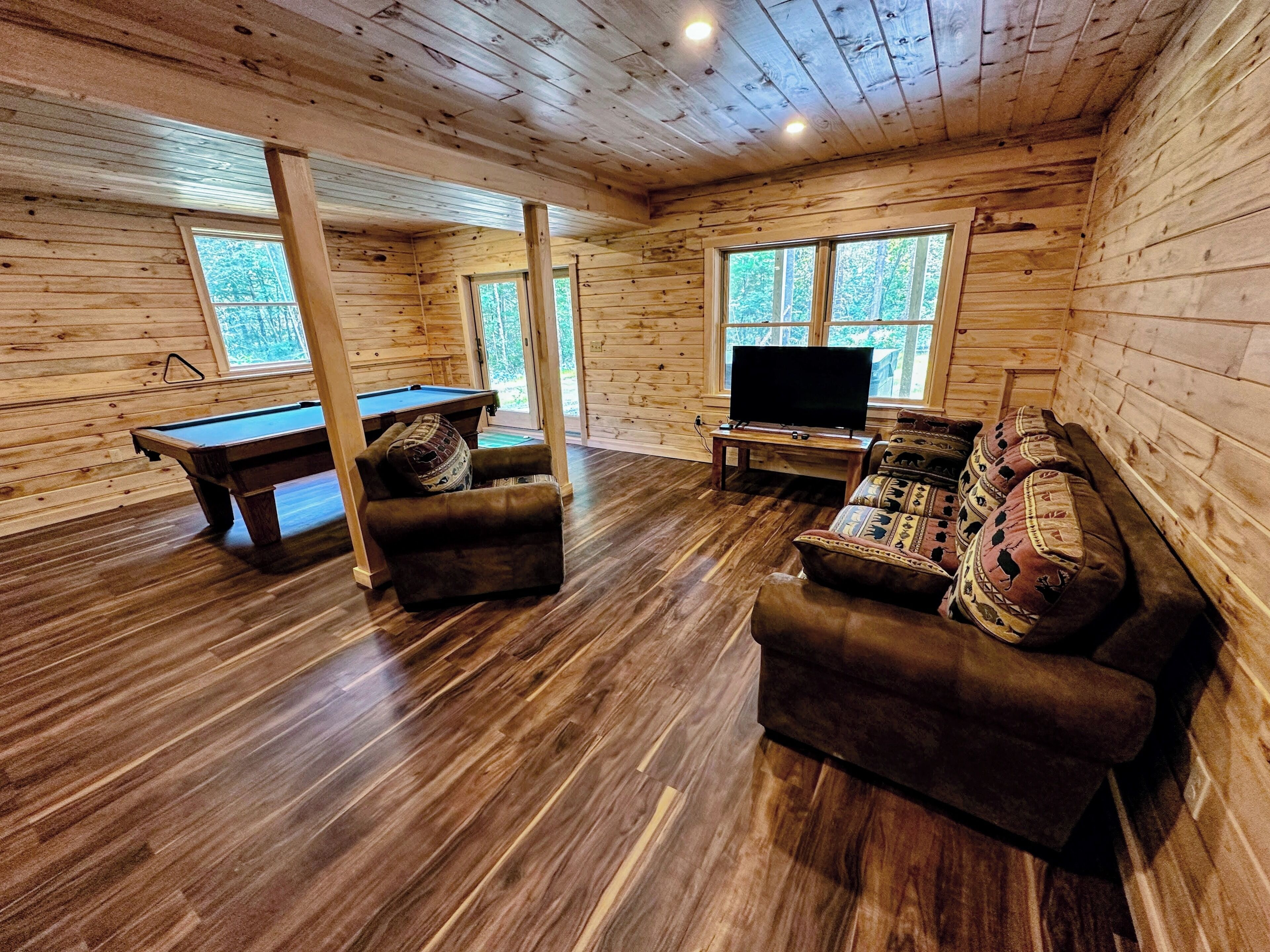 A wooden cabin interior with a pool table, a sofa, a television, and large windows.