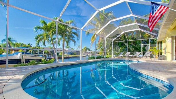 A swimming pool with a clear blue water, surrounded by a glass enclosure, palm trees, and a flag. - Villa Viscaya (Cape Coral)