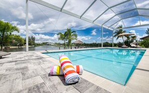 A swimming pool with a covered area, lounge chairs, and a view of a lake and palm trees. - Villa Little Diamond (Cape Coral)