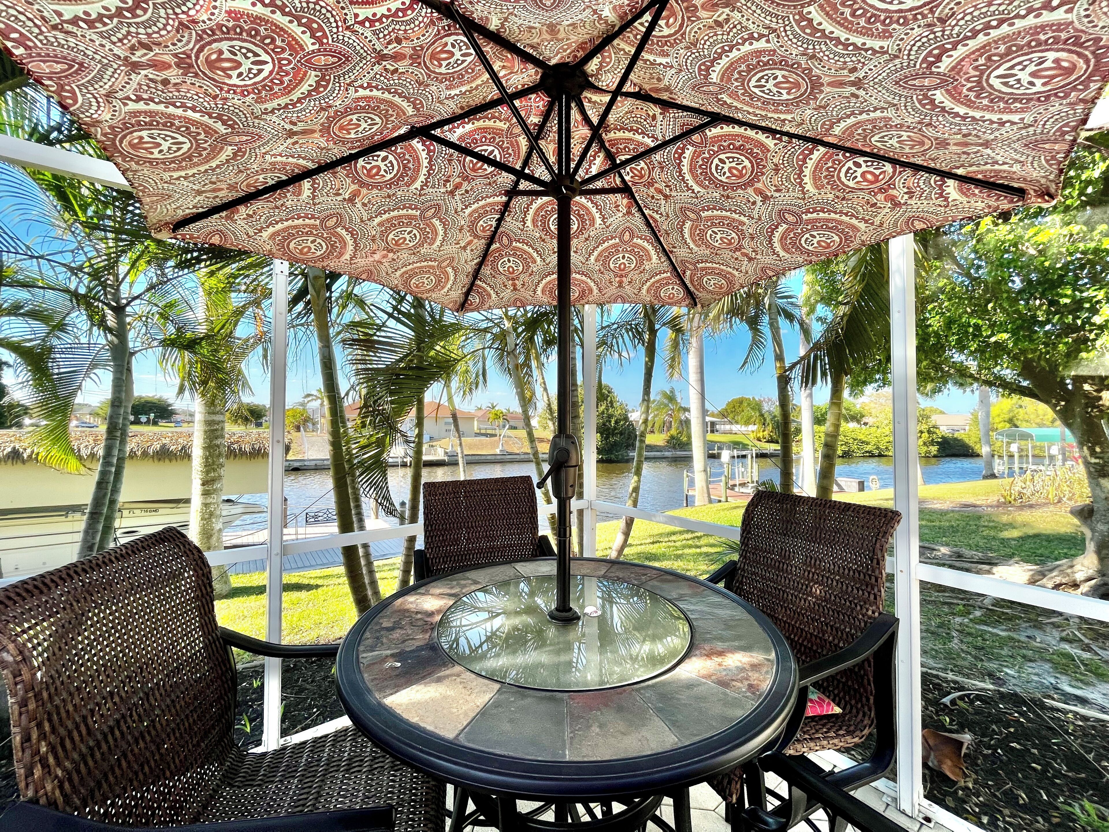 An outdoor seating area with a patterned umbrella, wicker chairs, and a glass table.