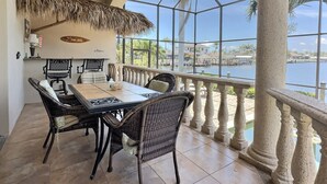 Ein Essbereich mit Tisch und Stühlen, einer reetgedeckten Decke und Blick auf ein Gewässer mit Gebäuden im Hintergrund.