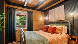 Ein Schlafzimmer einer Holzhütte mit einem Bett, einem Nachttisch, einem Fenster mit Vorhängen und einer Wandlampe.