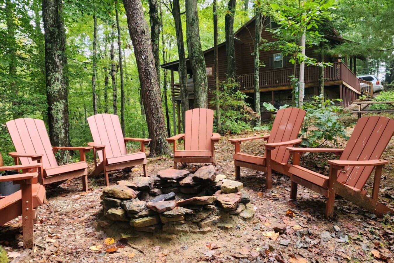 A cabin surrounded by a forest with a fire pit and four Adirondack chairs.