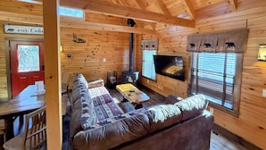 Ein Cabin-Innenraum mit Holzbalkendecke, Kamin, einer Couch, einem Fernseher und einem Essbereich.