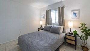 Une chambre à coucher avec un lit, deux tables de chevet avec des lampes, une fenêtre avec des rideaux et un tableau accroché au mur.