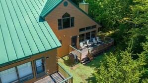 A two-story house with a green roof, a deck with outdoor seating, and a large window overlooking a forested area. - Mwp25 Updated 3 bed 3 Bath Condo With Mountain ski Slope Views in Bretton Woods (Bretton Woods)