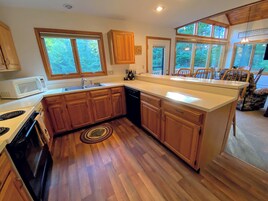Una cocina con armarios de madera, microondas, estufa y fregadero. Se ven árboles a través de las ventanas.