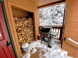 Una cabaña de madera con chimenea, leñera y un porche cubierto de nieve.