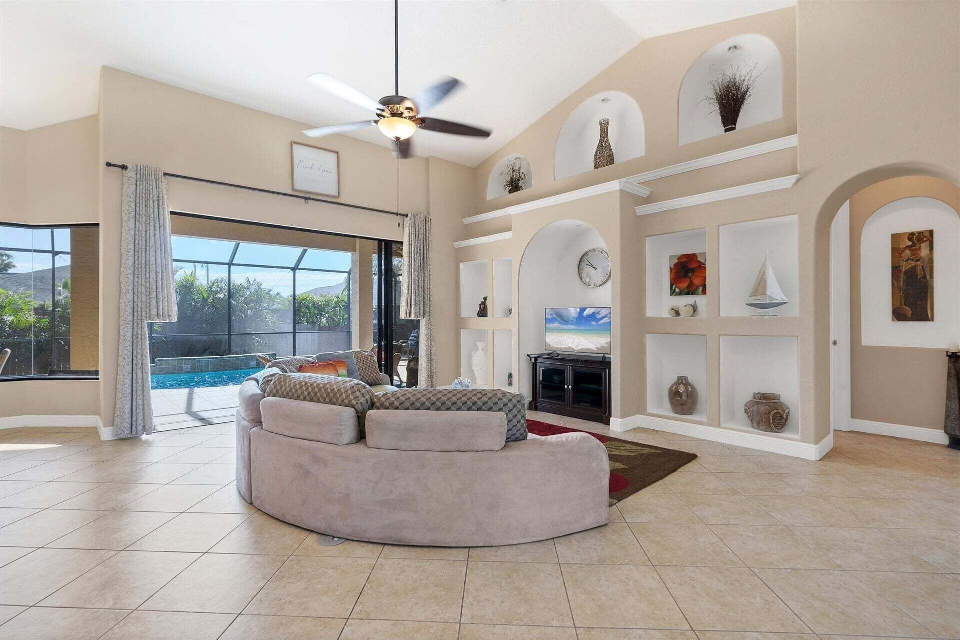 Un vaste salon doté d’un canapé incurvé, d’un ventilateur de plafond, d’un grand téléviseur et d’une piscine visible à travers la porte vitrée.