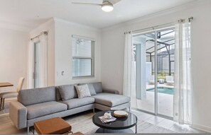 Una sala de estar moderna con un sofá seccional gris, una pequeña mesa redonda y un vidrio que da a un balcón con piscina.