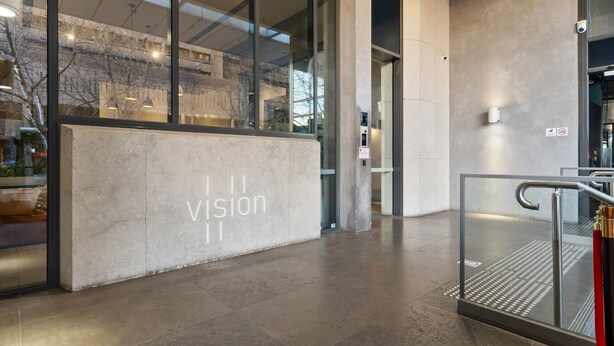 Una entrada de edificio moderna con fachada de vidrio, un mostrador de recepción de concreto y una escalera de metal.