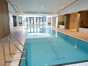 Ein Hallenbad mit deutlich abgegrenzten Bahnen, einem Rettungsschwimmerstuhl und Blick auf die Stadt durch große Fenster.