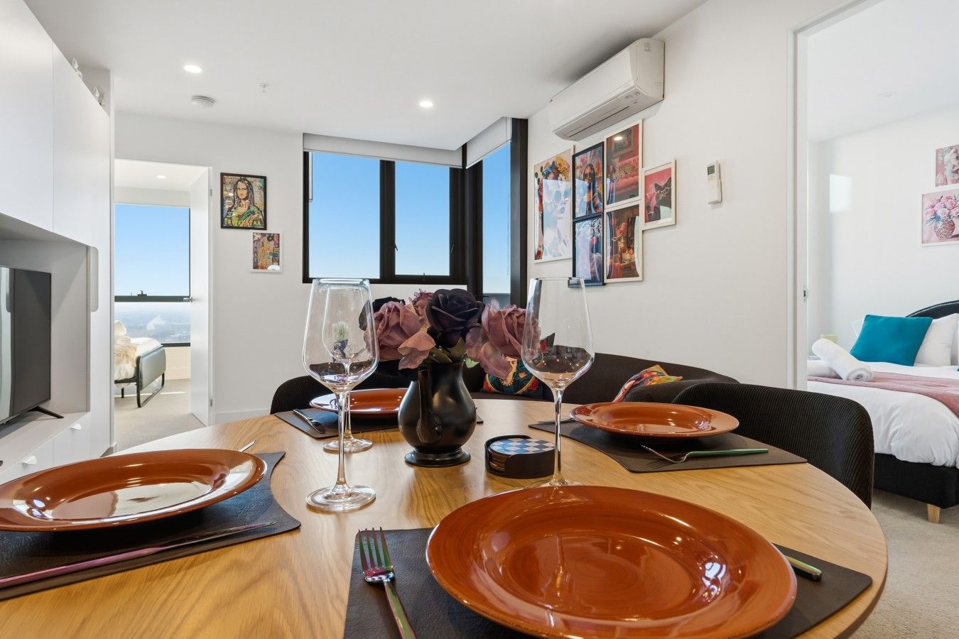 A modern hotel room with a dining area, a bed with a view, and artwork on the walls.