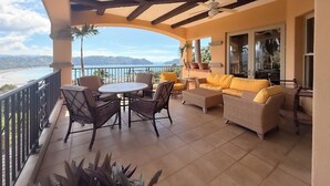 Een balkon met uitzicht op de zee, ingericht met rieten stoelen en een bank, en een plafondventilator.