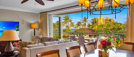 Habitación de hotel con balcón, ventilador de techo, televisor, zona de comedor con sillas y mesa, y vista a palmeras y edificios.