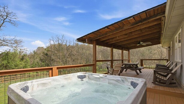A hot tub on a wooden deck with a covered area, chairs, and a view of trees. - Lazy Bear Meadow | Dog-Friendly Cabin with Fenced Yard, Hot Tub & Putting Green (Sevierville)