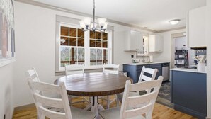 A dining area with a round table and white chairs, a kitchen with a blue island, and a large window with a view of trees. - Lazy Bear Meadow | Dog-Friendly Cabin with Fenced Yard, Hot Tub & Putting Green (Sevierville)