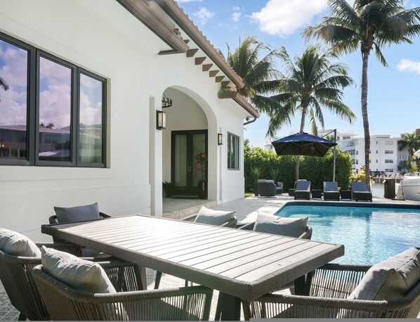 A poolside area with a wooden table and wicker chairs, a door leading to the house, and palm trees in the background. - Private Waterfront Luxury Villa Resort-style Living Privacy (Fort Lauderdale)