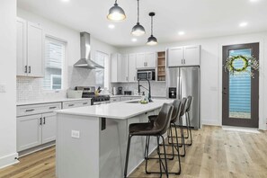 A modern kitchen with white cabinetry, a central island with bar stools, stainless steel appliances, and pendant lighting. - Near Indy Attractions • Deck • Patio • Fenced Yard (Indianapolis)