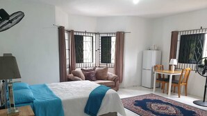 A bedroom with a bed, a desk, a chair, a refrigerator, and a fan.