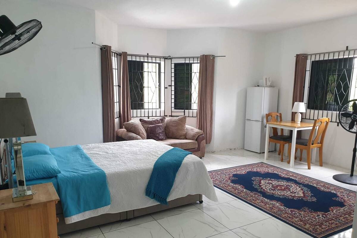 Un dormitorio con cama, escritorio, silla, refrigerador y ventilador.