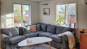 Un salon chaleureux avec un canapé d’angle gris, une table basse blanche et deux fenêtres ornées de plantes.