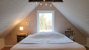 Une chambre mansardée chaleureuse avec un plafond en pente, une grande fenêtre, un lit recouvert d’une courtepointe blanche, une lampe de chevet et une petite table avec une lampe.
