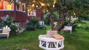 Une terrasse en bois blanc, agrémentée d’un banc rembourré et d’un hamac, entourée d’une végétation luxuriante et d’un bâtiment rouge doté de fenêtres.