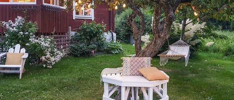 En hvid træterrasse med en polstret bænk og en hængekøje, omgivet af frodig vegetation og en rød bygning med vinduer.