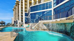 Area piscina di un resort con acqua cristallina, una cascata in pietra e un edificio a più piani con balconi.
