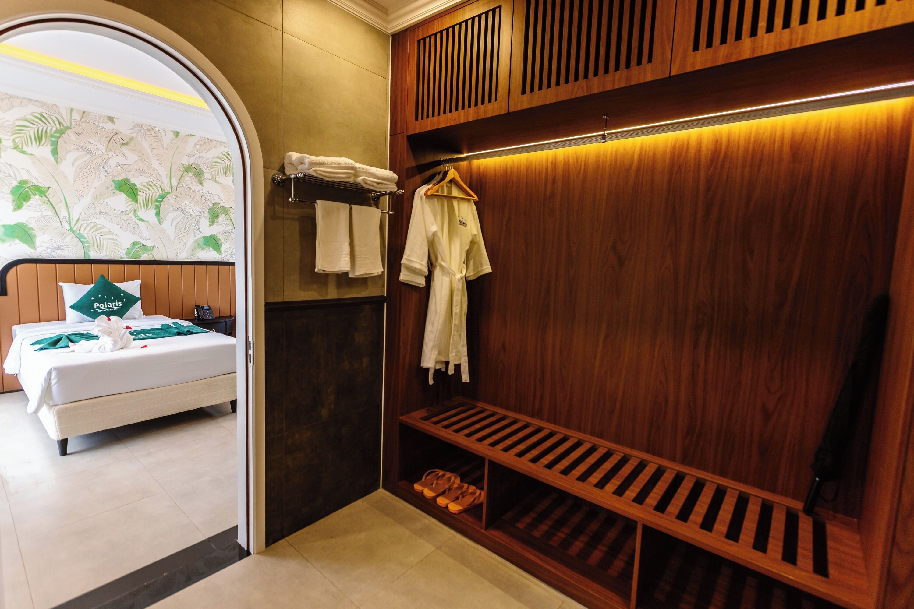 A hotel room with a bed, a wooden wardrobe, a shoe rack, and a towel rack.