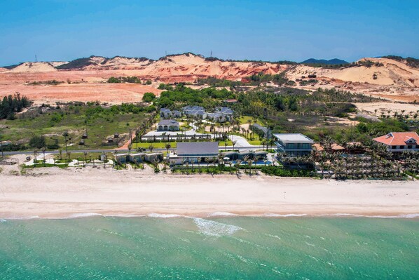 Ein Küstenresort mit Strandvillen, einem zentralen Gebäude, möglicherweise für Dienstleistungen, und einer hügeligen Kulisse.