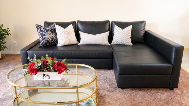A black leather sectional sofa with patterned cushions, a glass coffee table with a floral design, and a small sign with the word "joy".