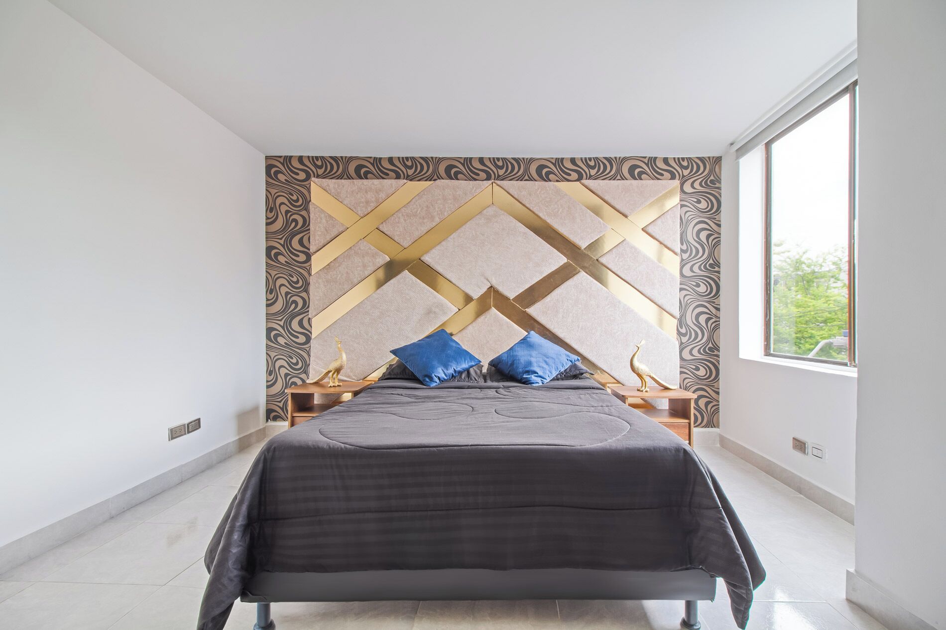 Een moderne slaapkamer met een hoofdeinde met een geometrisch patroon, een groot bed met een donkergrijze deken en twee blauwe kussens.