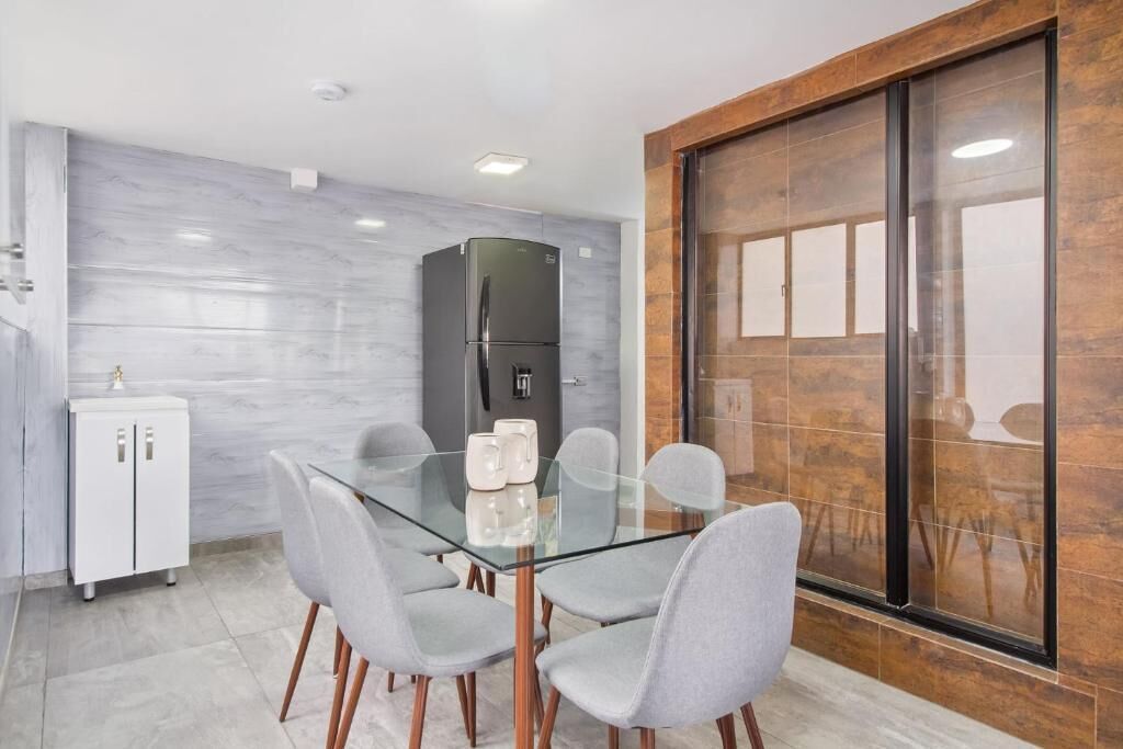 A modern kitchen with a glass dining table, grey chairs, a white cabinet, and a black refrigerator.
