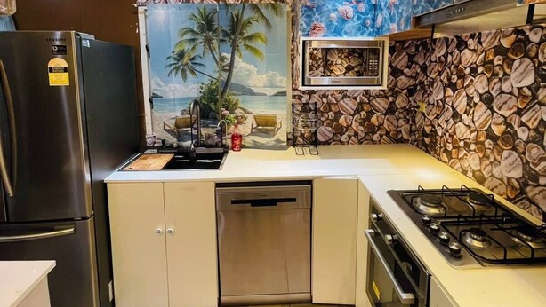 Une cuisine compacte équipée d’un réfrigérateur, d’une cuisinière, d’un four à micro-ondes et d’un lave-vaisselle. Les murs sont décorés d’une fresque tropicale et d’un motif représentant des coquillages.