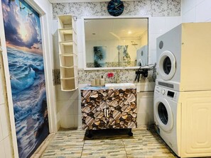 A compact bathroom with a tiled floor, a sink with a decorative backsplash, a mirror, a clock, and a washing machine. - Tropical Getaway in Nadi Town with Smart Home (Nadi Western Division)