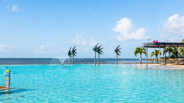 A clear blue pool with three windmill sculptures, a beach, and a covered area with palm trees. - Cairns Harbour Haven | Marina Views | Pool & Spa | near Reef Terminal (Cairns City)
