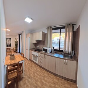 A compact kitchen with a microwave, stove, and sink, wooden cabinets, and a dining area with a table and chairs. - Holiday Apartment Gara (Adeje)