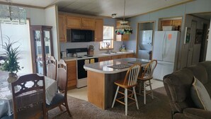 A compact kitchen with wooden cabinets, a microwave, a refrigerator, and a dining area with a table and chairs. - Cozy 2 bedroom Lake Cottage on Lamoka Lake in Bradford New York (Bradford)